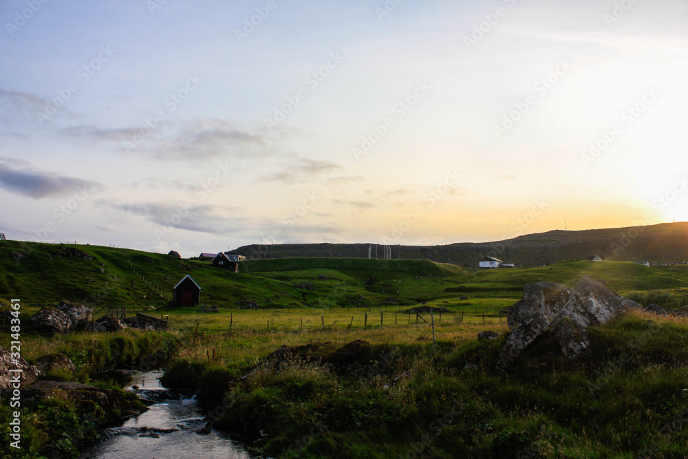 Fototapeta premium Green hilly meadow and small houses on it. Evening sunset. Sunlight.