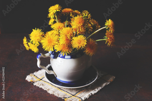 Bouquet of Bright Yellow Dandellions in A White And Blue Teapot Standing On the Blue Saucer On the Napkin photo