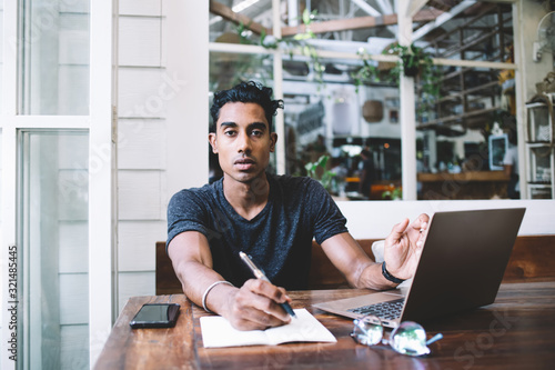 Concentrated ethnic student taking notes in notebook while surfing laptop in cafe