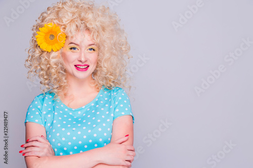 Beautiful blonde girl with voluminous curly hairstyle  in a blue polka dot blouse and with a flower of a sunflower in her hair on a gray background