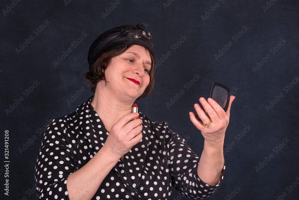 Waist up portrait of an elderly retro smiling woman wearing hat looks in the mirror and paints her lips with lipstick against a dark background with copy space Waist up portrait of an elderly retro smiling woman wearing hat looks in the mirror and paints her lips with lipstick against a dark background with copy space