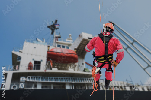 Safety man worker Abseiling, rope access sprinkle from structure steel with safety harness and wearing equipment protective PPE floating dock in shipyard on blue sky background