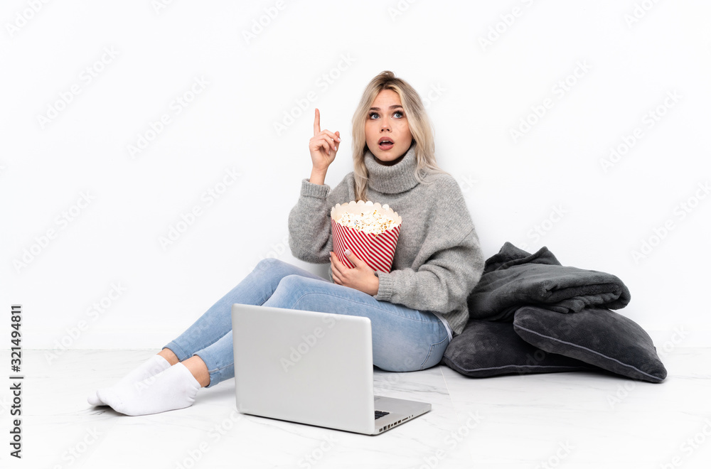 Teenager blonde girl eating popcorn while watching a movie on the laptop intending to realizes the solution while lifting a finger up Teenager blonde girl eating popcorn while watching a movie on the laptop intending to realizes the solution while lifting a finger up