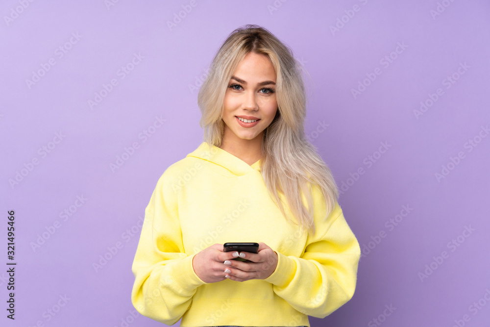 Teenager girl wearing a yellow sweatshirt over isolated purple background sending a message with the mobile Teenager girl wearing a yellow sweatshirt over isolated purple background sending a message with the mobile