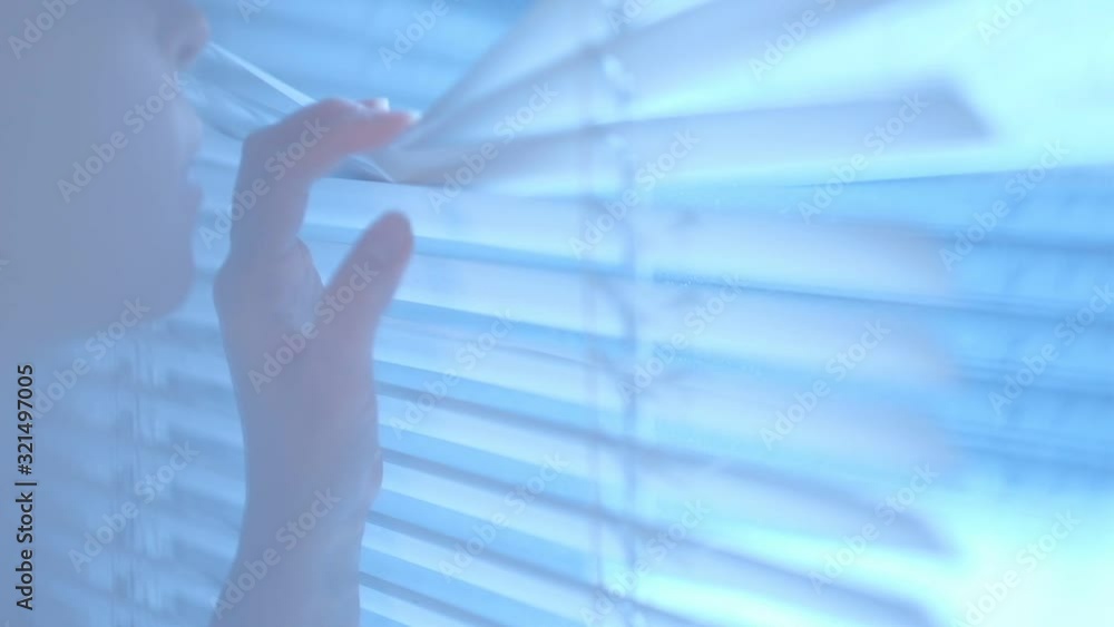Intrigued pretty young woman looks out the window through the blinds then closes them by passing them by her hand. Close up of a hand reaching and opening a crack in the window blinds Intrigued pretty young woman looks out the window through the blinds then closes them by passing them by her hand. Close up of a hand reaching and opening a crack in the window blinds