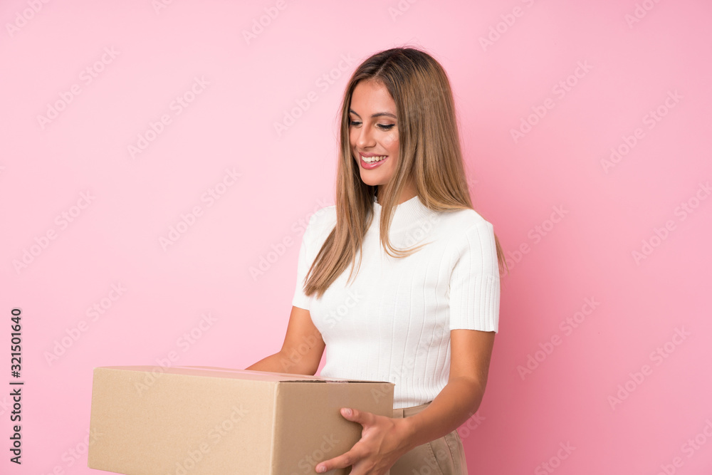 Young blonde woman over isolated pink background holding a box to move it to another site Young blonde woman over isolated pink background holding a box to move it to another site