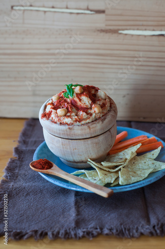 Wooden bowl with chickpea hummus and red paprika on a blue plate with carrot sticks and nachos with a rustic background of a brown and wooden handkerchief photo