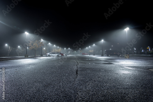 A Lonely Parking Lot on a Cold, Rainy Night - with a Small Collection of Parked Cars in the Background and a Thick Mist Gathering Under the Lights