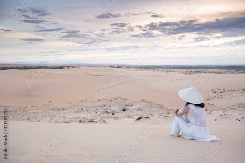 Female in white gown and traditional Vietnamese conical hat sitting on the sand dunes of Mui Ne on sunset with copy space, Mui Ne, Vietnam