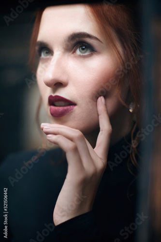 Portrait of a red-haired girl in a black turtleneck and trousers with professional makeup from a stylist. A shot near a vintage window.