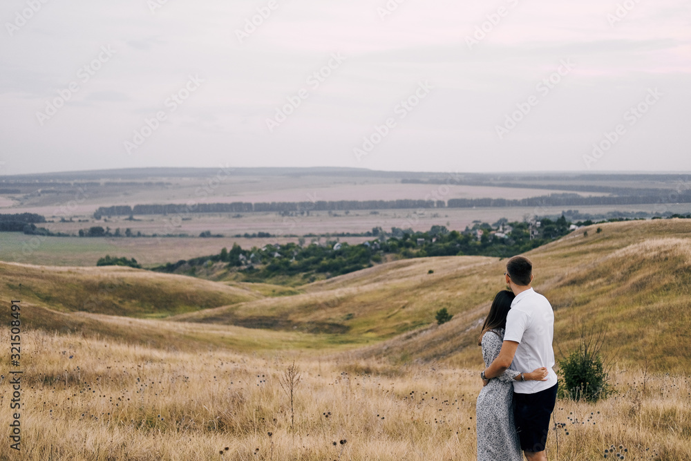 A young couple in love stands in a field on a hill and looks into the distance. Hug and admire the summer landscape.