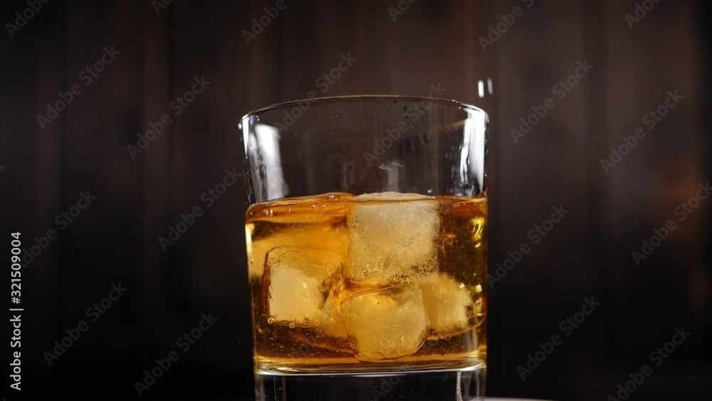 Close-up of ice cubes falling into a rotating glass of whiskey on a dark wooden background, a lot of spray flying in different strons. Slow motion. Artificial lighting. Close-up of ice cubes falling into a rotating glass of whiskey on a dark wooden background, a lot of spray flying in different strons. Slow motion. Artificial lighting.