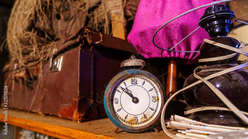 Old rusty alarm clock stands on a wooden shelf on the background of junk. Timeless and garage sale concept.