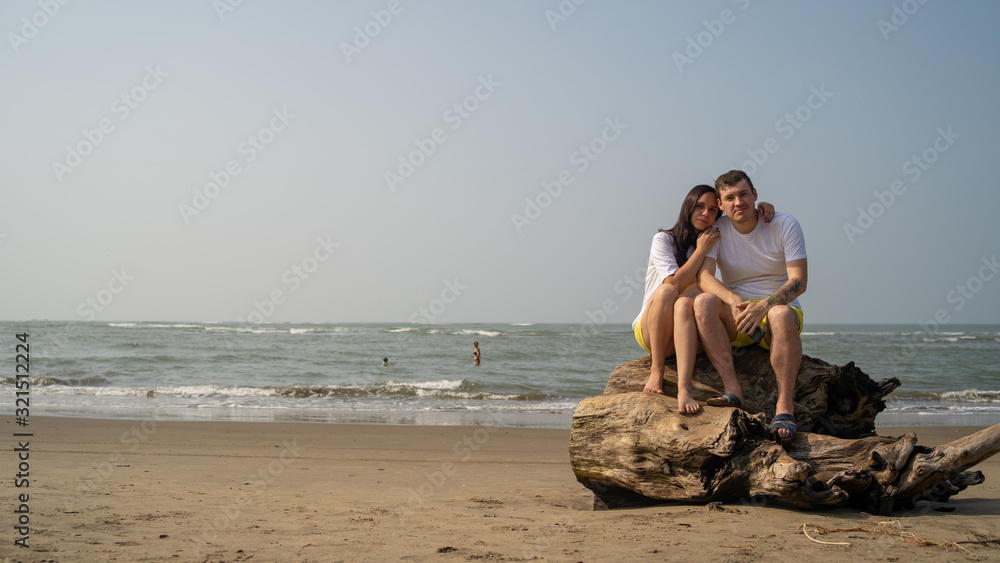 Happy couple posing on driftwood near sea. Loving couple hugging during date on beach against waving sea and cloudless sky.