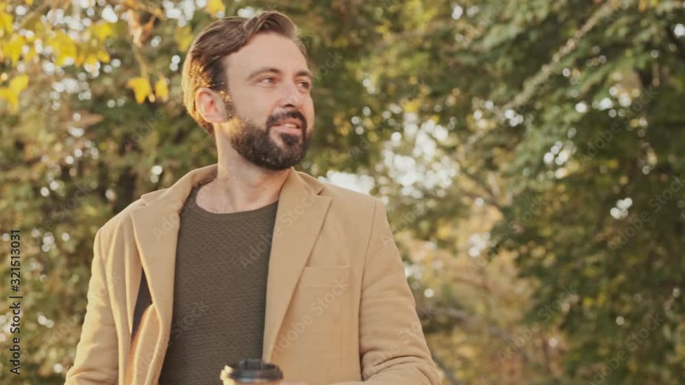 Smiling elegant bearded man in coat walking with cup of coffee in the park outdoors Smiling elegant bearded man in coat walking with cup of coffee in the park outdoors