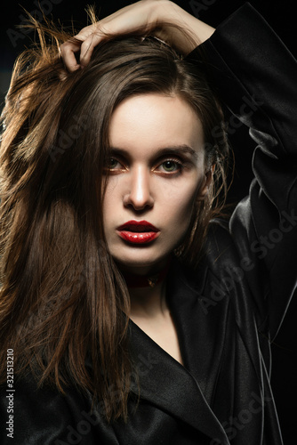 Beautiful slim girl model with red lips  wearing a black blazer  touches her hair with her hand on a dark background  posing next to a light lamp. Advertising  trendy  and lifestyle design.