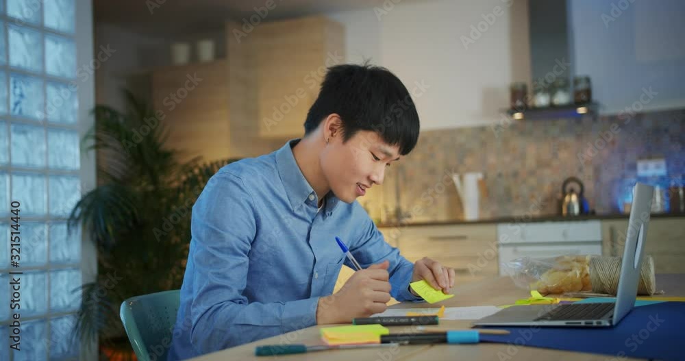 Attractive young Asian creative man writing notes on colorful stickers alone at home, freelance artist designer enjoying work in front of laptop dressed in blue shirt smiling indoor modern kitchen Attractive young Asian creative man writing notes on colorful stickers alone at home, freelance artist designer enjoying work in front of laptop dressed in blue shirt smiling indoor modern kitchen