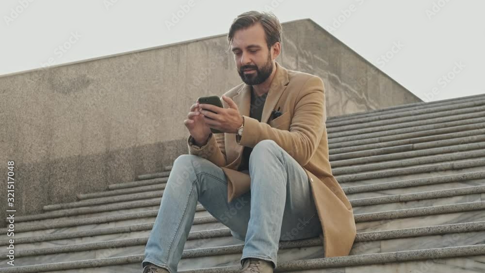View from below of smiling elegant bearded man in coat using smartphone and looking away while sitting on stairs outdoors View from below of smiling elegant bearded man in coat using smartphone and looking away while sitting on stairs outdoors