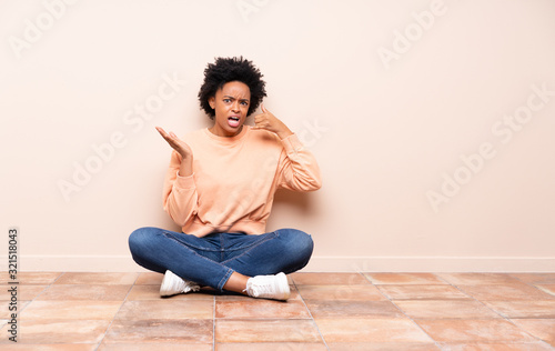 African american woman sitting on the floor making phone gesture and doubting