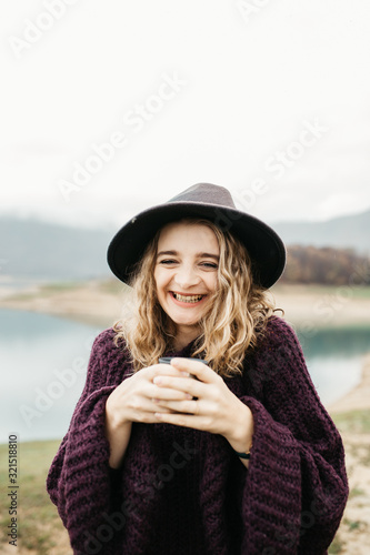 Happy beautiful woman with hat drinking coffee in nature, on a foggy morning. Lake and mountains are in the background. She is holding cup of coffee.