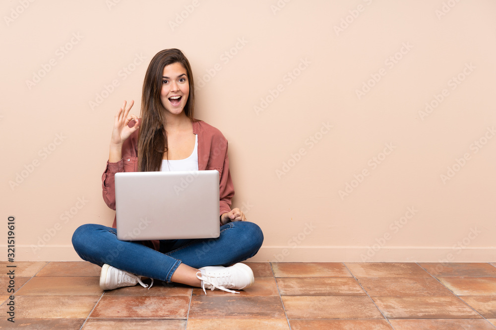 Teenager student girl sitting on the floor with a laptop surprised and showing ok sign Teenager student girl sitting on the floor with a laptop surprised and showing ok sign
