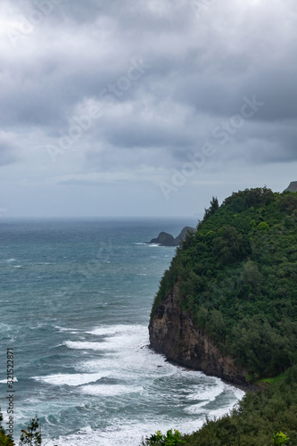 Kohala, Hawaii, USA. - January 15, 2020: Portrait of Coastline where Pololu valley and its beach meets the ocean. Big rocks, tall steep cliffs, green forests under heavy blueish cloudscape.