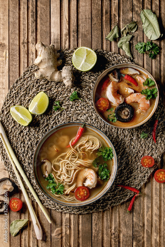 Variety of asian soups. Traditional spicy Thai tom yum kung and noodles soup with shiitake mushrooms, prawns, ingredients above on straw wicker napkin over wooden plank background. Flat lay, space.