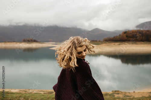 happy girl with curly blond hair dances on a lake alone, her hair is flying because of the wind flow, free as a bird. photo of girl with curly hair standing back to camera over lake background.