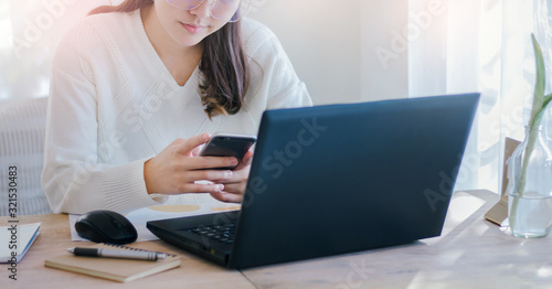 Young female using mobile phone to searching information while she working with laptop on desk