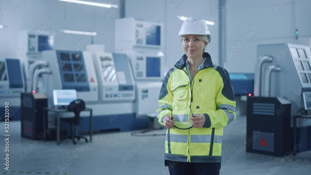 Beautiful Smiling Female Engineer Wearing Safety Vest and Hardhat Takes of Safety Goggles. Professional Woman Working in the Modern Manufacturing Factory. Facility with CNC Machinery and robot arm Beautiful Smiling Female Engineer Wearing Safety Vest and Hardhat Takes of Safety Goggles. Professional Woman Working in the Modern Manufacturing Factory. Facility with CNC Machinery and robot arm