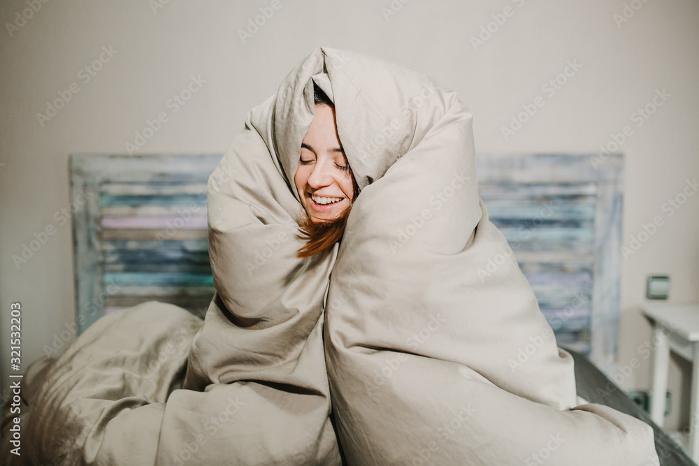 Fototapeta premium .Pretty young woman smiling just woken up wrapped in her large quilt on a cold winter morning. Lifestyle.