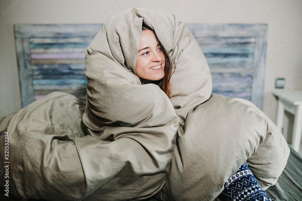 Fototapeta premium .Pretty young woman smiling just woken up wrapped in her large quilt on a cold winter morning. Lifestyle.