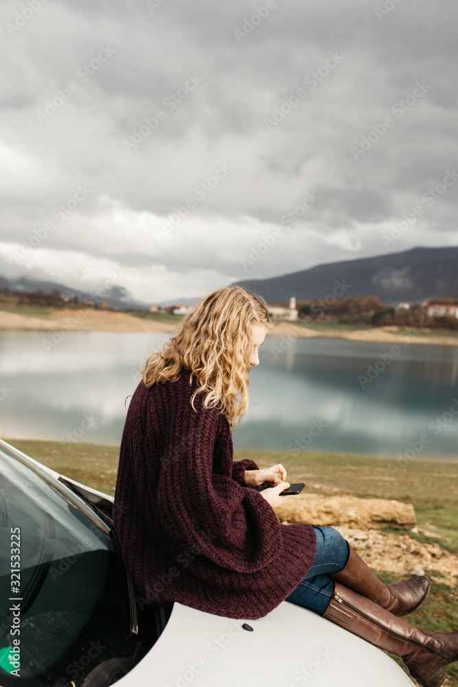 Beautiful woman with curly hair, sitting on the car, holding mobile phone and taking photos of lake on a cloudy day. She is texting on smartphone and taking selfie. 