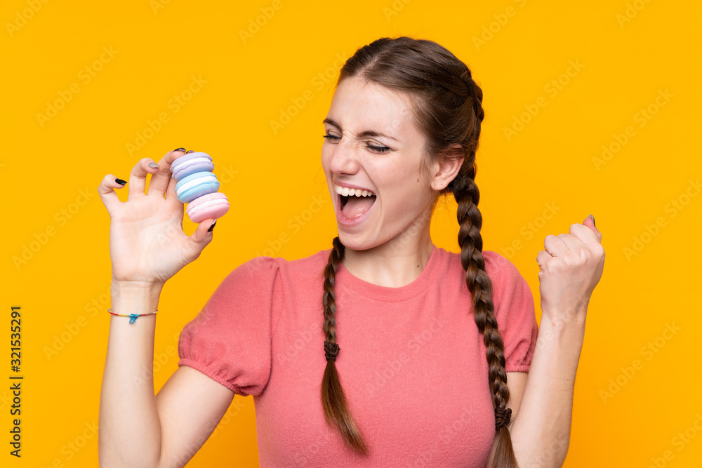 Young woman over isolated yellow background holding colorful French macarons and celebrating a victory