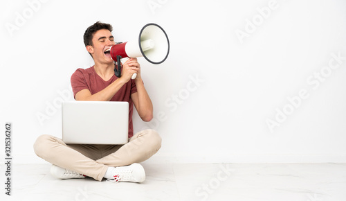 Teenager man sitting on the flor with his laptop shouting through a megaphone