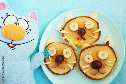 A plate with three pancakes in the shape of cat faces, decorated with slices of banana, grape and apple and a soft toy cat with an orange nose. Children's food serving. Art food...