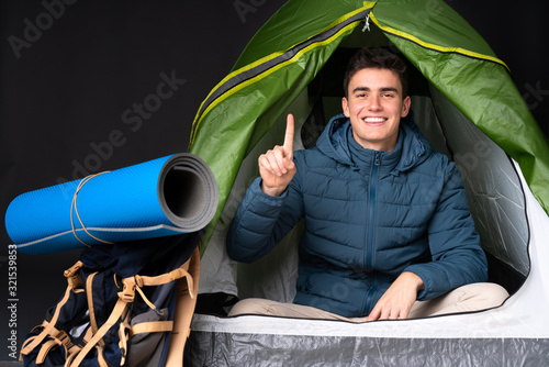 Teenager caucasian man inside a camping green tent isolated on black background showing and lifting a finger in sign of the best