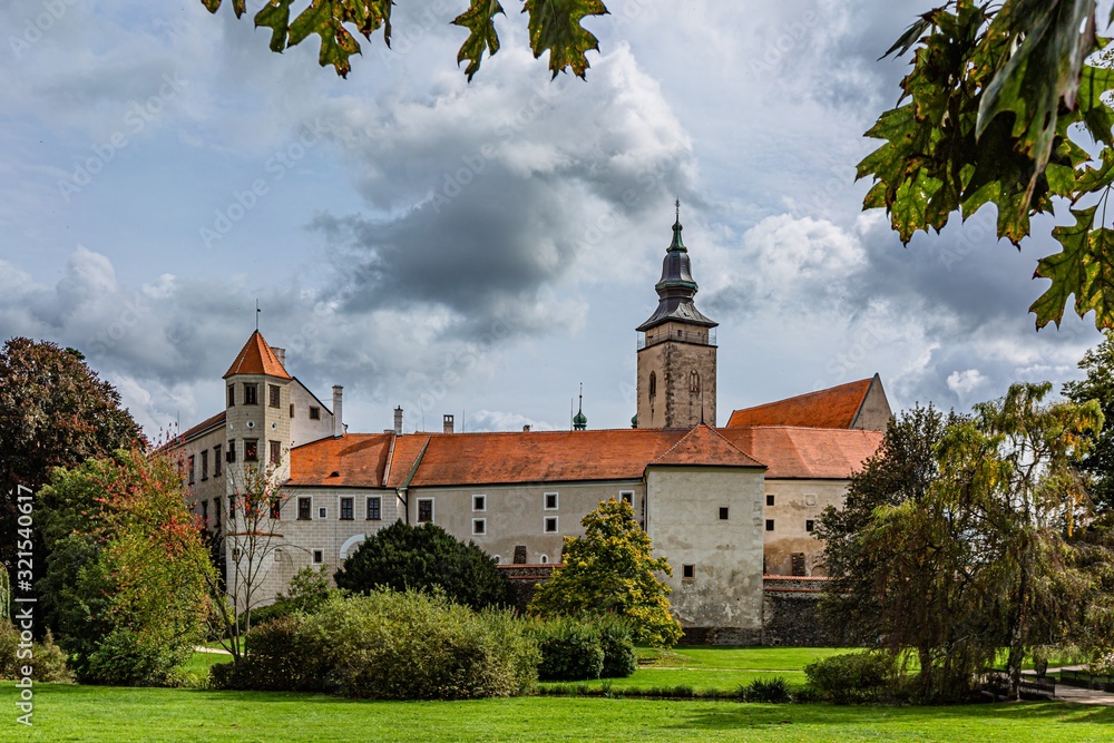 Telc / Czech Republic - September 27 2019: View of a state castle and a tower of the church of James the Great from a public park with green trees and grass. Sunny day with blue clouds.
