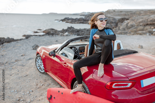 Portrait of a young woman surfer in swimsuit sitting with surfboard on the red cabriolet on the rocky ocean coast. Active lifestyle and surfing concept