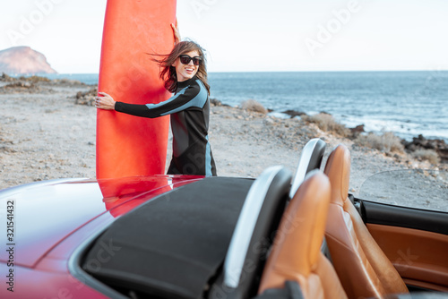 Portrait of a young woman surfer in swimsuit standing with surfboard behind her red car on the rocky coast