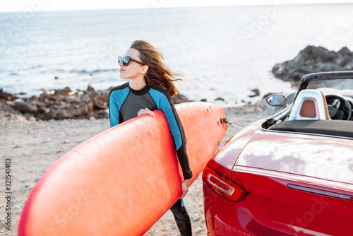 Photography Portrait of a young woman surfer in swimsuit standing with surfboard near the car on the rocky coast