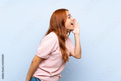 Teenager redhead girl over isolated blue background shouting with mouth wide open to the lateral