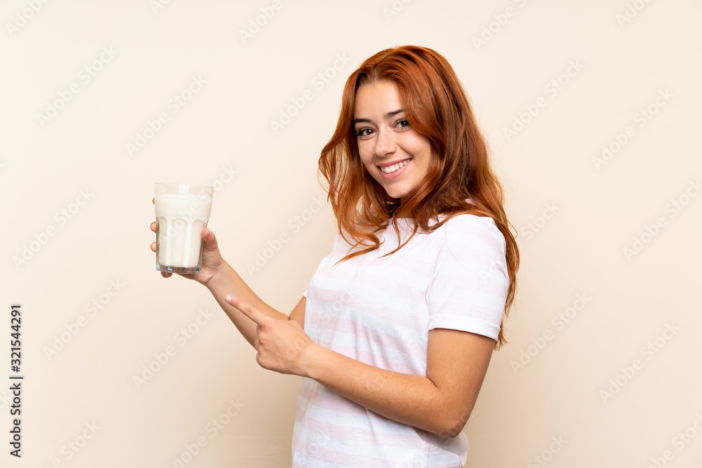 Teenager redhead girl holding a glass of milk over isolated background and pointing it Teenager redhead girl holding a glass of milk over isolated background and pointing it