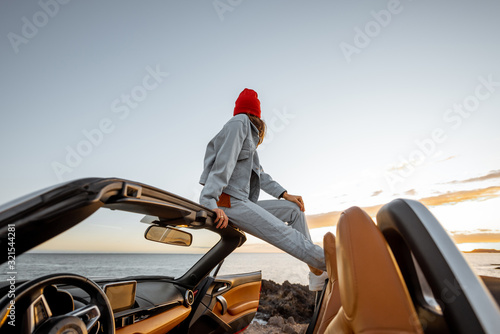 Woman enjoying beautiful view on the ocean, sitting on the car roof top during a sunset. Nature enjoyment and carefree travel by car concept