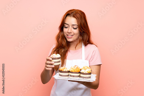 Teenager redhead girl holding lots of different mini cakes over isolated pink background