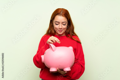 Teenager redhead girl with sweater over isolated green background holding a big piggybank