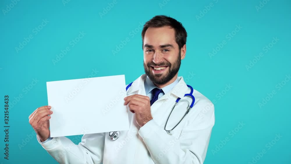 Portrait of young handsome man in professional medical white coat holding white horizontal a4 paper isolated on blue studio background. Doctor with beard and stethoscope. Copy space. Portrait of young handsome man in professional medical white coat holding white horizontal a4 paper isolated on blue studio background. Doctor with beard and stethoscope. Copy space.