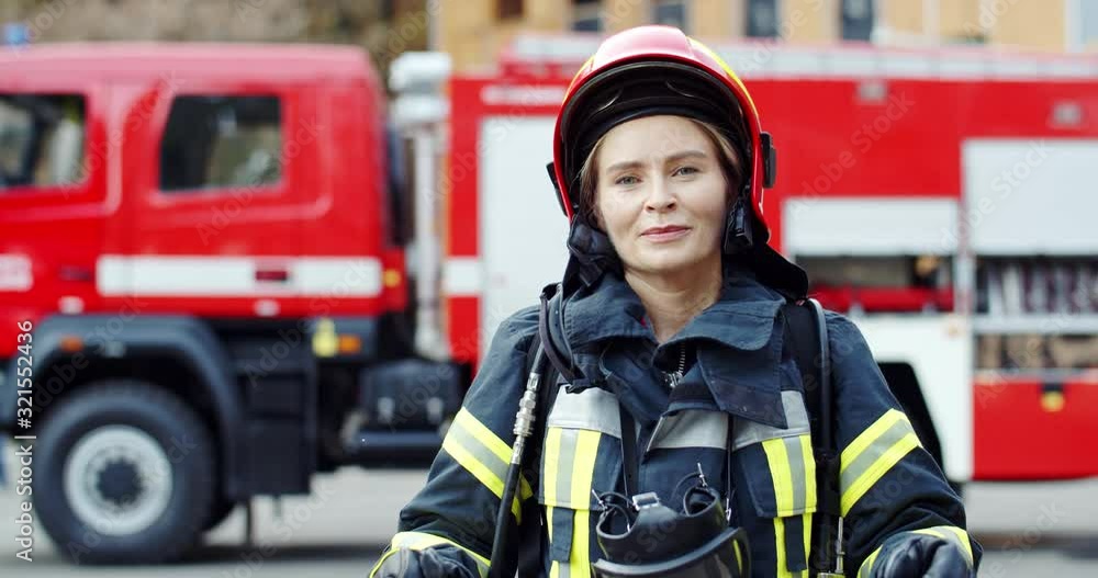 Portrait shot of the pretty and happy Caucasian woman firefighter in the special costume and helmet taking off goggles and smiling joyfully to the camera. Outdoors. Portrait shot of the pretty and happy Caucasian woman firefighter in the special costume and helmet taking off goggles and smiling joyfully to the camera. Outdoors.