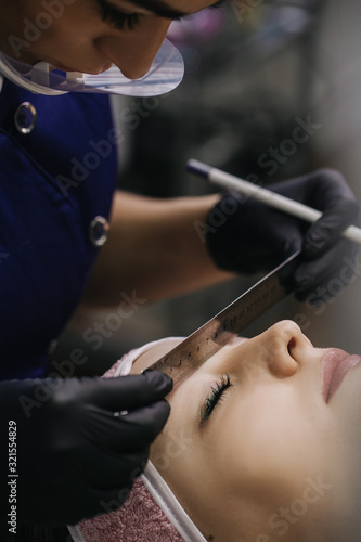 The master in a protective mask in black gloves holds a metal ruler over the eyebrows of a girl who has her eyes closed and looks at the model’s eyebrows.