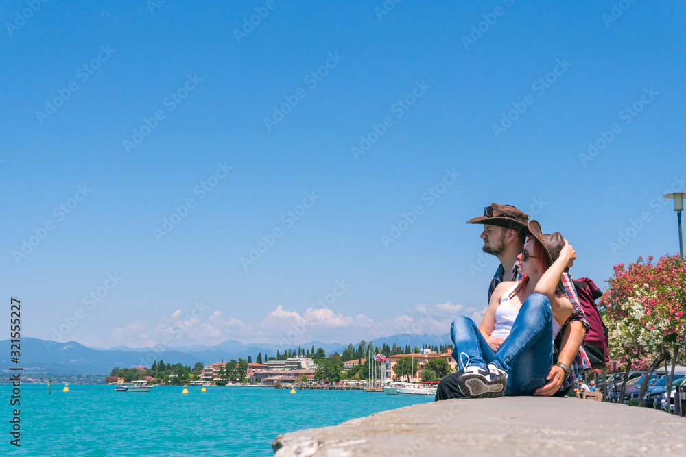 A couple in a cowboy hat and a backpack sits on the promenade, looks at the turquoise water of Lake Garda. Vacation and tours to Europe. Sunny summer day. Italy. Mountains background, copy space